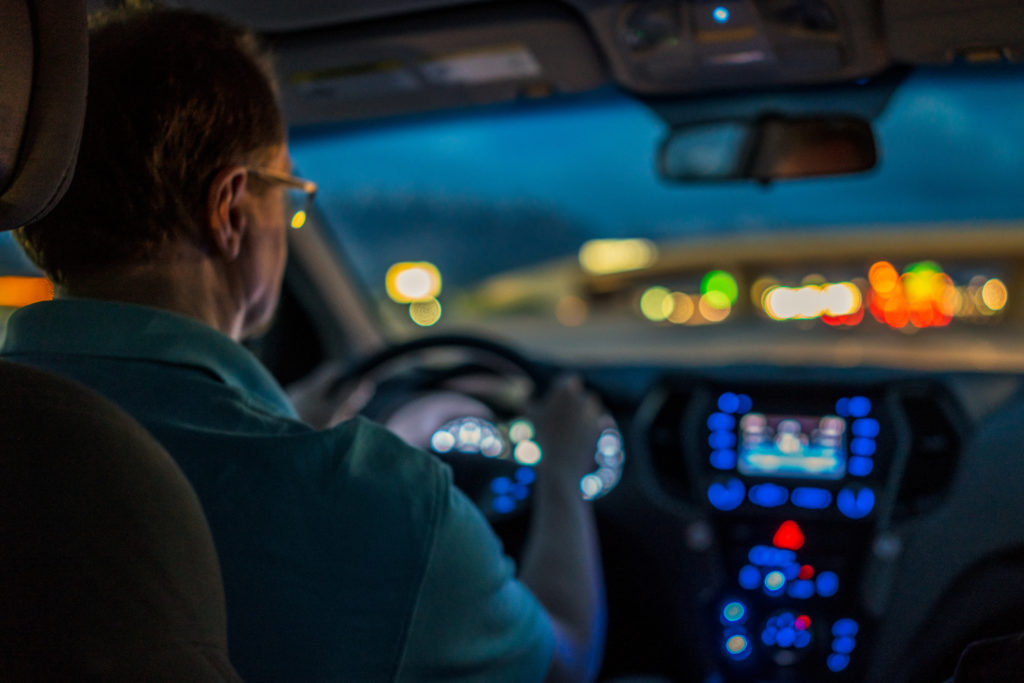 Passenger view of a man driving an SUV on a freeway in Utah, USA at night, with the freeway lights in the distance and the car's dashboard lights in the foreground. Shallow depth of field.