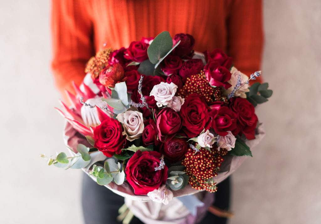 Very nice young woman in red sweater holding blossoming flower bouquet of fresh roses, carnations, eucalyptus in vivid red passionate colors on the grey wall background