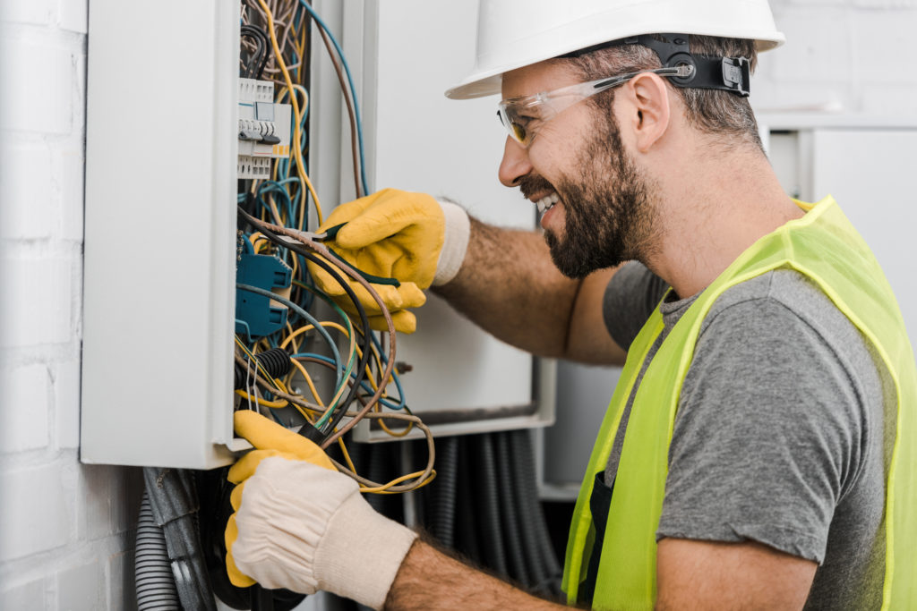 side view of smiling handsome electrician repairing electrical box with pliers in corridor electrician contractor insurance