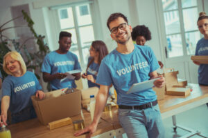 Man in volunteer shirt smiles while holding clipboard, group of volunteers behind him