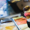 scooping ice cream on a cone at an ice cream shop; case full of brightly colored flavor options