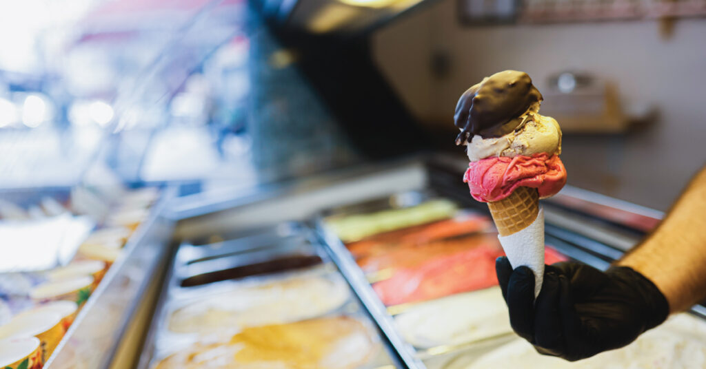 scooping ice cream on a cone at an ice cream shop; case full of brightly colored flavor options